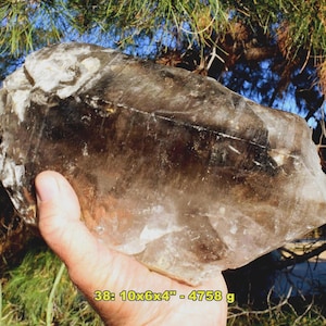 May include: A large, rough-hewn smoky quartz crystal, held in a hand. The crystal is a translucent brown color with areas of white and dark brown. The text on the image reads: 38: 10x6x4" - 4758 g.