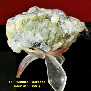 May include: A Prehnite mineral specimen from Morocco, displayed on a clear stand. The stone is a mix of pale green and white, with a crystalline structure. The text on the image reads "15: Prehnite - Morocco 2.5x1x1" - 100 g."