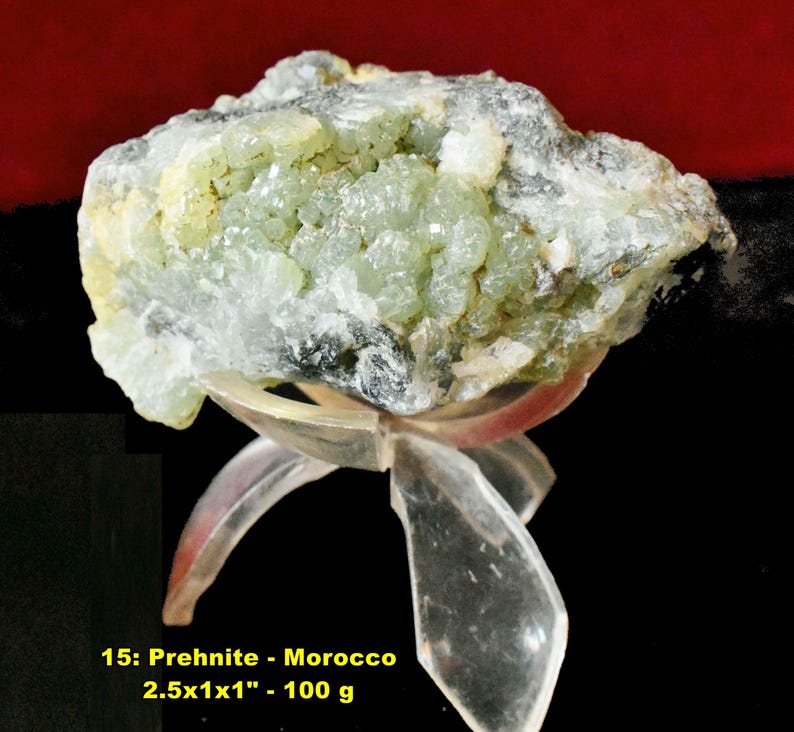 May include: A Prehnite mineral specimen from Morocco, displayed on a clear stand. The stone is a mix of pale green and white, with a crystalline structure. The text on the image reads "15: Prehnite - Morocco 2.5x1x1" - 100 g."