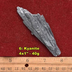 May include: A gray kyanite crystal, approximately 4 inches long, rests on a red surface. The text "6: Kyanite 4x1" - 40g" is visible. A wooden ruler is in the foreground.