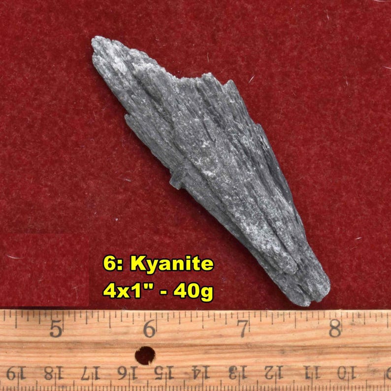 May include: A gray kyanite crystal, approximately 4 inches long, rests on a red surface. The text "6: Kyanite 4x1" - 40g" is visible. A wooden ruler is in the foreground.