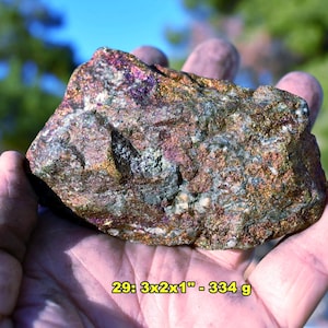 May include: A raw, multi-colored mineral specimen featuring metallic copper, gold, and purple iridescent hues with a rough, crystalline texture. The rock is labeled with the text 29: 3x2x1" - 334 g, indicating its dimensions and weight.