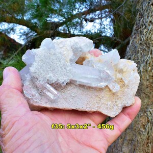 May include: A cluster of clear quartz crystals with a light pink hue, set on a beige matrix. The specimen is approximately 5 x 3 x 2 inches and weighs 456 grams. The crystals are held in a hand, with a natural background.