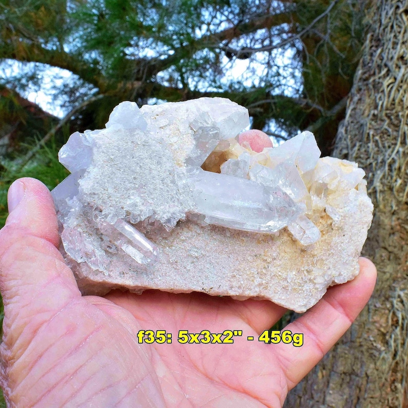 May include: A cluster of clear quartz crystals with a light pink hue, set on a beige matrix. The specimen is approximately 5 x 3 x 2 inches and weighs 456 grams. The crystals are held in a hand, with a natural background.