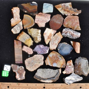 May include: A collection of various polished stones and minerals in shades of brown, beige, gray, and purple. The stones have unique patterns and textures. Two dice and a ruler are also present.