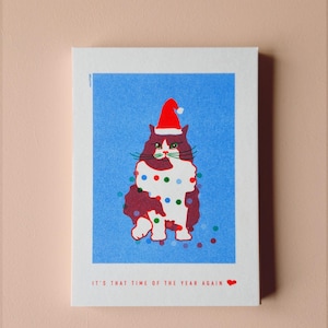 May include: A greeting card featuring a whimsical illustration of a cat wearing a Santa hat and Christmas lights. The cat is depicted in shades of burgundy and white against a blue background. The text "IT'S THAT TIME OF THE YEAR AGAIN" is printed at the bottom.