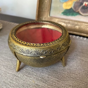 May include: A gold-toned trinket box with a glass lid and three legs. The box has a red velvet interior and intricate engraved details.