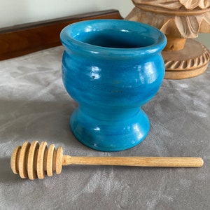 May include: A blue ceramic honey pot with a wooden honey dipper. The honey pot has a rounded body and a flared lip. The honey dipper is made of wood and has a spiral design.