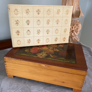May include: A wooden box with a floral design on the lid. The box is closed and has a light brown finish. The lid is decorated with a pattern of gold fleurs-de-lis.