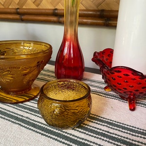 May include: A set of four vintage glass bowls. Two amber bowls with a textured surface, one with a floral design and one with a round shape. A red bowl with a textured surface and three legs. A red glass vase with a flared top.