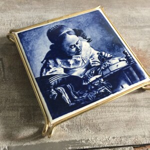 Puede incluir: Un azulejo de cerámica azul y blanco con un marco de metal dorado que representa a una niña con un vestido blanco con detalles azules, sentada a una mesa y trabajando en un trozo de tela.