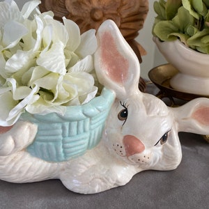 Puede incluir: Un macetero de cerámica blanca en forma de conejo con una cesta azul claro que contiene flores blancas. El conejo tiene orejas rosas y una cara pintada.