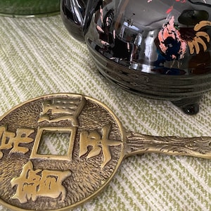 Könnte beinhalten: Ein chinesischer Glücksbringer aus Messing mit einem quadratischen Loch in der Mitte und chinesischen Schriftzeichen auf der Vorderseite. Der Glücksbringer hat einen Griff auf der Rückseite.