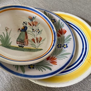Może przedstawiać: Trzy ceramiczne talerze z niebieskimi i żółtymi obwódkami i wzorami kwiatowymi. Najmniejszy talerz przedstawia kobietę w niebieskiej sukience i czerwonej spódnicy stojącą na polu kwiatów.