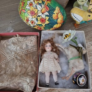 Puede incluir: Una muñeca de porcelana con cabello rubio y un vestido blanco está sentada en una caja con un conejo blanco y una flor. La muñeca tiene ojos azules y un rubor rosado en las mejillas. El conejo lleva una cinta verde alrededor del cuello.