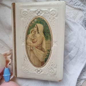 May include: Vintage white book featuring an oval image of a woman holding a child. The image is framed with a gold border and surrounded by ornate, embossed detailing. The book's spine is light brown.