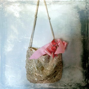 May include: A vintage-style beige crocheted handbag with a pink satin ribbon and floral embellishments. The bag has a long, woven strap and a delicate, openwork design. The interior is a lighter shade of beige.