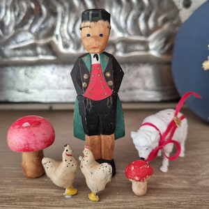 Puede incluir: Colección de pequeñas figuras de madera pintadas a mano. El conjunto incluye una figura con vestimenta tradicional, dos gallinas, una oveja con una cinta roja y dos setas. Las figuras están sobre una superficie de madera.