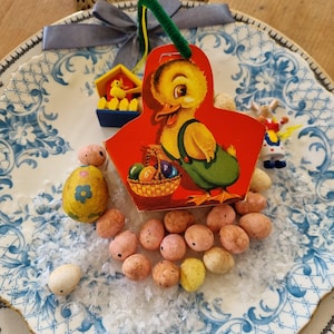 Cesta vintage de cartón para Pascua llena de huevos de espuma moteada con una mini casa de pájaro de madera alemana colgando de su asa.