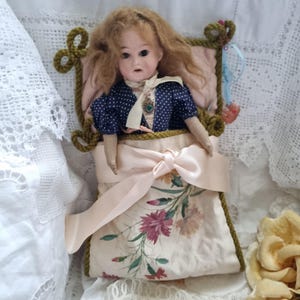 Könnte beinhalten: Eine Vintage-Puppe mit blondem Haar und einem blauen Polka-Dot-Kleid ruht in einem floral bedruckten Bett. Das Bett hat ein rosa Kissen und ist mit einem hellrosa Band gebunden. Die Puppe hat eine Halskette und ist vor einem weißen Spitzengrund platziert.