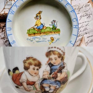 May include: A vintage porcelain children's dish and teacup set. The dish features a cartoon rabbit fishing with a mouse. The teacup has illustrations of two children with a rabbit. The dish has blue lettering around the rim.