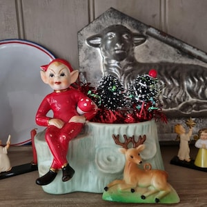 May include: A vintage Christmas ornament featuring a red elf figurine seated on a light blue planter with miniature Christmas trees and a deer figurine. The planter has a tree stump design. A metal lamb relief is in the background.