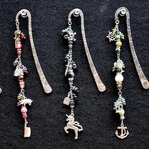 May include: Six silver bookmark pendants with various charms and beads. The charms include a cat, a pig, a unicorn, a skull, a ship, a palm tree, an anchor, and a compass. The beads are in shades of red, green, and brown.