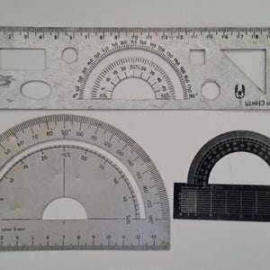 Peut inclure: Un ensemble de trois outils de dessin comprenant une règle métallique rectangulaire avec des découpes géométriques, un grand rapporteur métallique semi-circulaire gradué et un petit rapporteur semi-circulaire noir avec une base quadrillée.