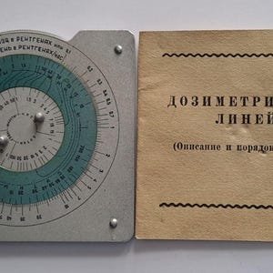 Può includere: Un regolo calcolatore dosimetrico quadrato in metallo grigio con un quadrante circolare rotante color verde acqua con scale numeriche e testo in cirillico, accompagnato da un manuale cartaceo color marrone chiaro intitolato "ДОЗИМЕТРИЧЕСКАЯ ЛИНЕЙКА".