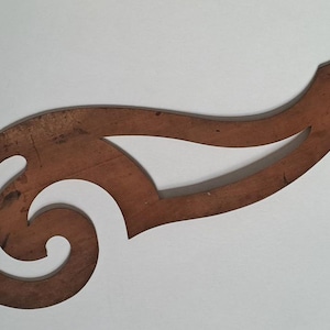 May include: A brown wooden French curve drafting tool featuring a smooth, flowing, asymmetrical shape with a central cutout. The surface of the wood shows natural grain patterns and minor signs of wear, with a pointed end and a curved base.