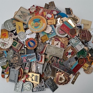 Può includere: Una vasta collezione di spille e distintivi in metallo vintage assortiti, caratterizzati da forme, dimensioni e colori vari. Gli oggetti presentano diversi design grafici, tra cui testo in cirillico, simboli nazionali, monumenti e ritratti.