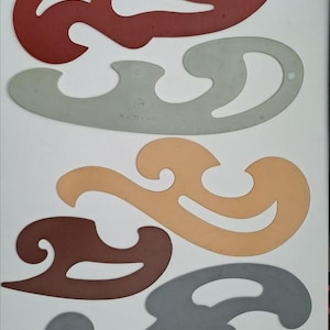 Puede incluir: Un conjunto de cinco plantillas de curvas francesas en varios colores, incluyendo rojo, gris claro, beige, marrón y gris oscuro. Estas herramientas de dibujo planas de plástico presentan bordes curvos, irregulares y suaves para trazar formas.