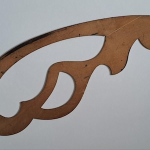 Può includere: Un curvilinee da disegno in plastica color marrone chiaro semitrasparente. Lo strumento presenta una forma complessa e irregolare con molteplici bordi curvi e ritagli, mostrando segni di usura con piccoli graffi e segni sulla superficie.
