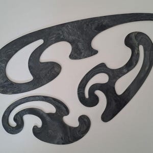 May include: A set of three dark gray French curve drafting templates featuring a marbled pattern. These drafting tools have various irregular, smooth, and curved edges designed for drawing complex shapes and lines on paper surfaces.