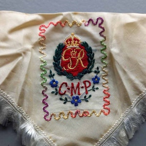 Puede incluir: Un pañuelo de seda blanco con un borde de flecos. El pañuelo presenta un diseño bordado con una corona, las letras "C.M.P." y un motivo floral. El bordado está hecho en hilo rojo, azul, verde y amarillo.