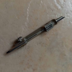 Può includere: Un compasso argentato con una punta nera e una manopola sul lato. Il compasso è appoggiato su una superficie marrone chiaro.