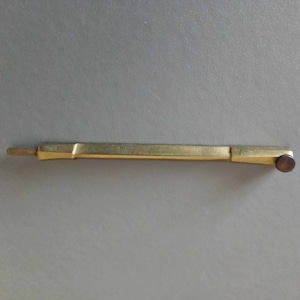 Peut inclure: Un objet rectangulaire de couleur laiton avec un petit bouton rond brun foncé à une extrémité. L'objet a un design légèrement incliné et semble être en métal. L'article est posé sur un fond gris.