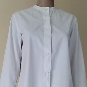 Peut inclure: Une chemise blanche à manches longues avec un col officier et une fermeture boutonnée. Les manches sont aux trois quarts. La chemise est présentée sur un mannequin.