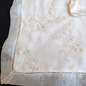 Könnte beinhalten: Elfenbeinfarbenes Leinentextil mit zarter Blumenstickerei. Die Stickerei zeigt kleine rosa und weiße Blüten mit grünen Stielen und Blättern. Die Kanten sind mit einer gewellten Bordüre versehen.