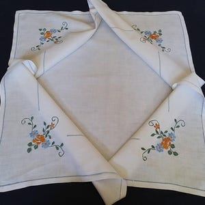 Può includere: Tovaglia di lino bianco con ricami floreali in arancio, blu e verde. La tovaglia è piegata a metà, mostrando il ricamo su entrambi i lati.