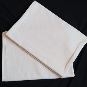 Op de afbeelding: Twee gevouwen, gebroken witte lakens op een zwarte achtergrond. De lakens zijn gemaakt van een zachte, geweven stof. Het bovenste laken is diagonaal gevouwen, waardoor de rand van het onderste laken zichtbaar is. De lakens lijken nieuw.