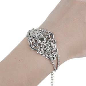 Op de afbeelding: Een zilveren armband met een groot, sierlijk filigraanontwerp. De armband heeft een delicate ketting die zich uitstrekt vanaf het hoofdontwerp.