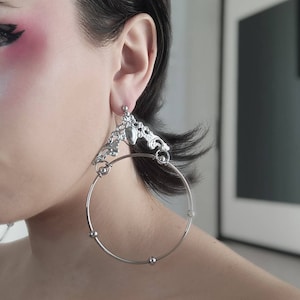 Può includere: Orecchini a cerchio d'argento con un designo decorativo e ornato. L'anello è grande e ha un ciondolo d'argento appeso.