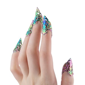 May include: A hand with long, iridescent, pointed nail covers. The nail covers have a decorative, openwork design and a metallic, multi-coloured finish. The hand is positioned against a white background.