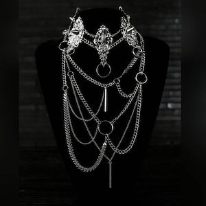 May include: A silver layered chain necklace with multiple chain styles, including a delicate chain, a curb chain, and a chain with small spikes. The necklace features several silver rings and a long silver bar pendant.