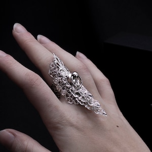 May include: A silver-colored statement ring with an intricate, openwork design. The ring features a large, oval-shaped gemstone and extends along the finger. The ring is displayed against a dark background.