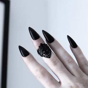 May include: A hand with long, black, pointed nails is wearing a black ring with a delicate, intricate design. The ring is on the middle finger.