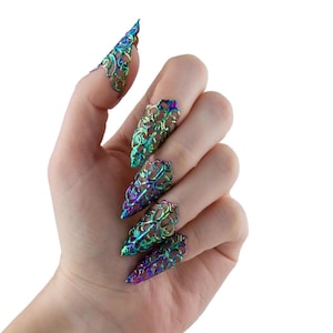 Pode incluir: Uma mão mostrando cinco capas de unhas pontiagudas iridescentes. As capas de unhas são uma mistura de verde, azul e roxo, com um design vazado. A mão está em um fundo branco.