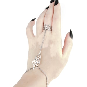 May include: A silver spiderweb ring with a chain that extends to the wrist, forming a bracelet. The ring and bracelet are both detailed with a spiderweb design.