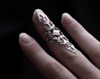 May include: A silver filigree ring with a delicate design, worn on the index finger of a hand.